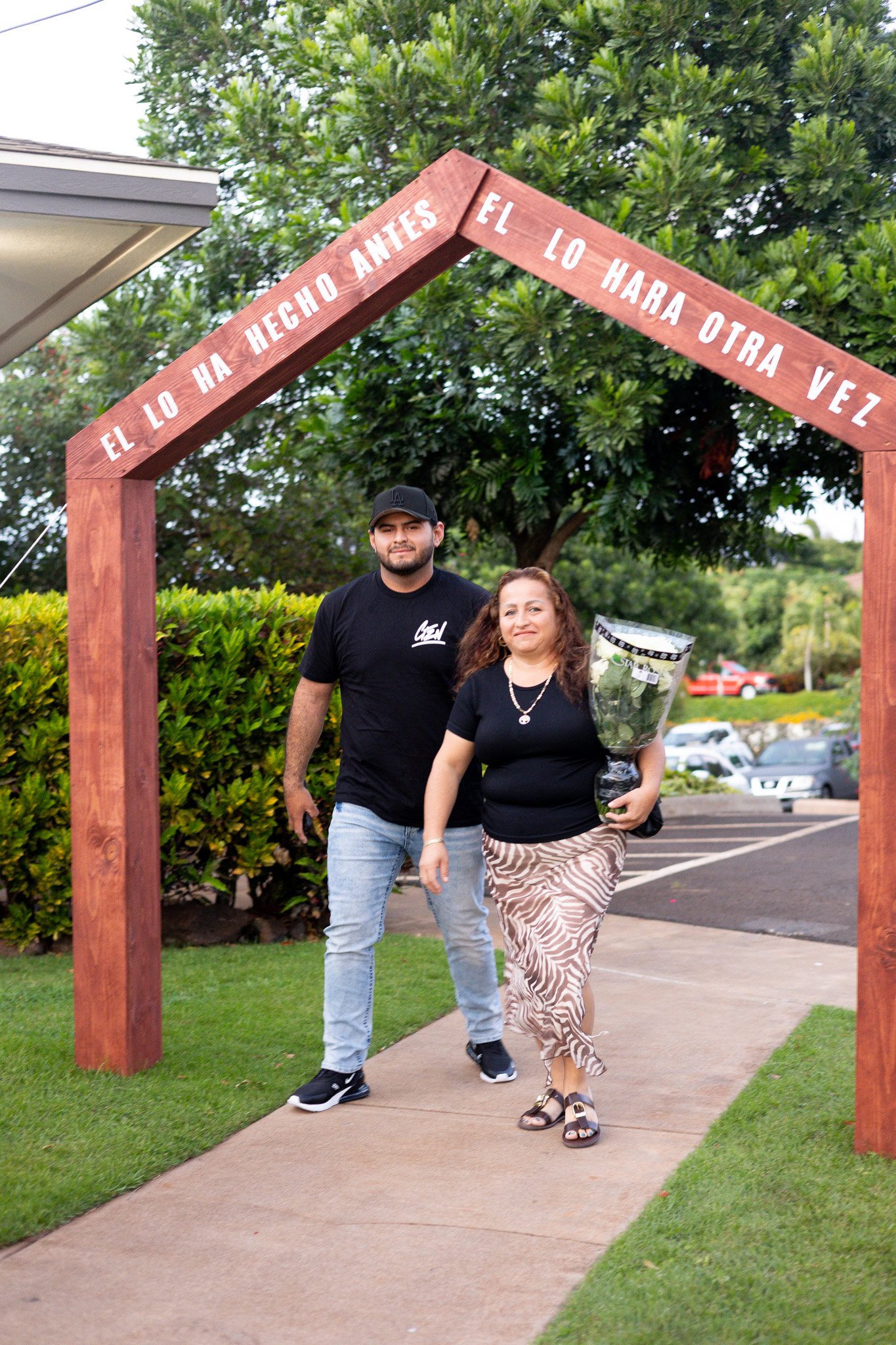 A woman walking on a sidewalk holding a bouquet of flowers, accompanied by a man, under a wooden archway that reads 'El lo hecho antes el lo har a otra vez.'