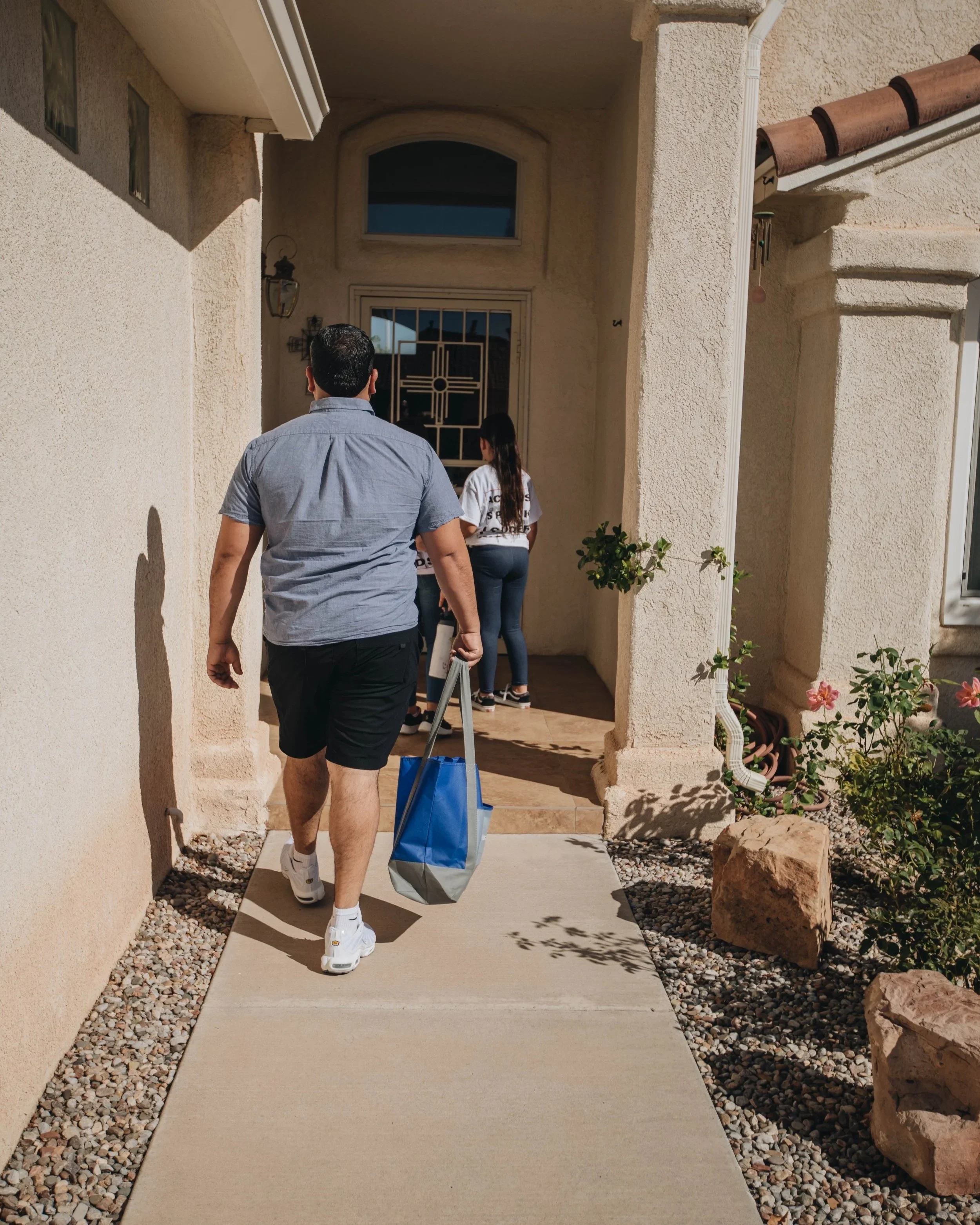 Man carrying a shopping bag walking toward a house entrance where two people are waiting inside.