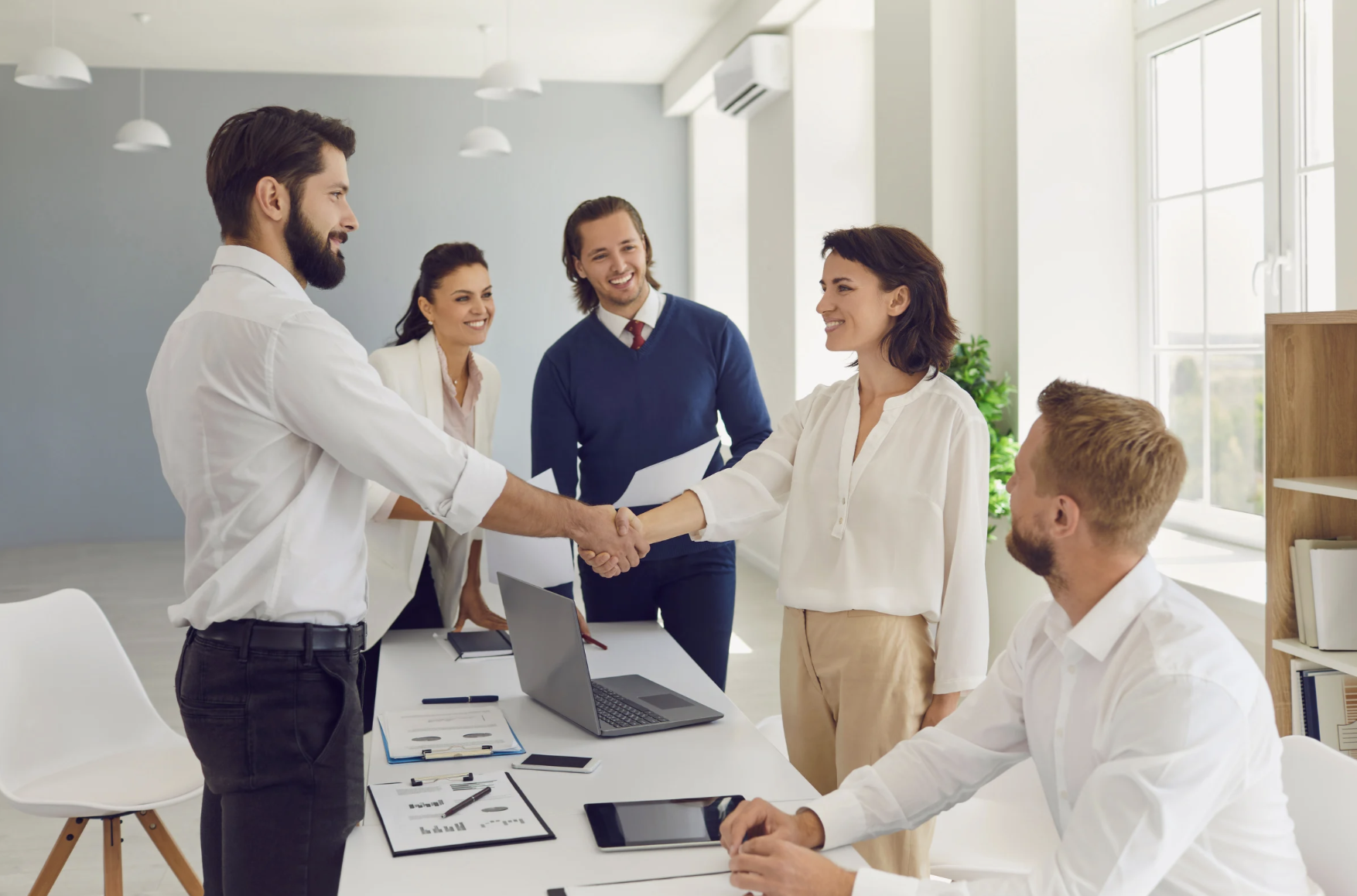 A group of five professionals in a bright office, with two people shaking hands, smiling. The scene shows a meeting or greeting, with laptops, documents, and phone on the table.