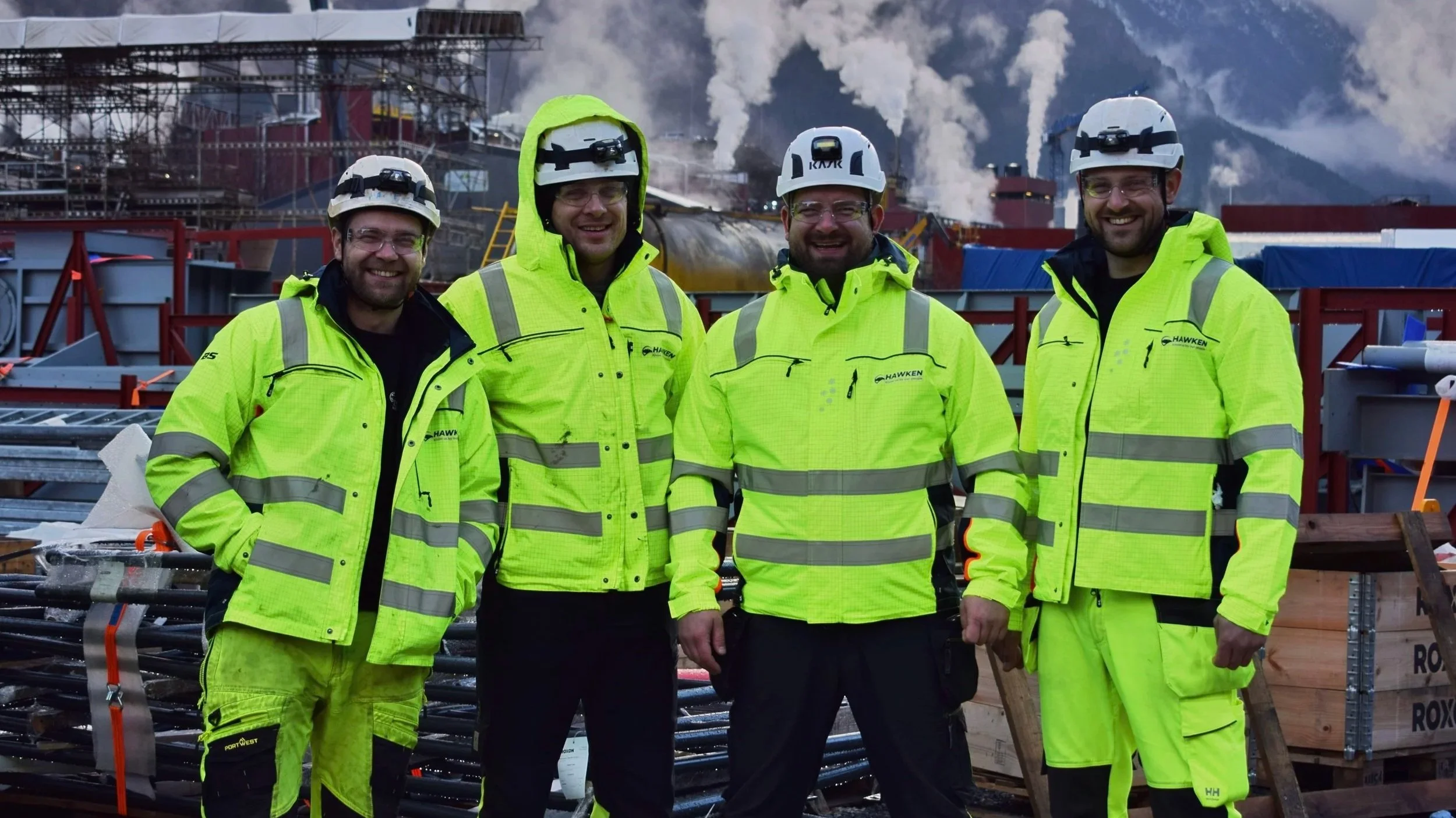 Fire arbeidere i høye, gule sikkerhetsvester og hjelm står foran industrielle anlegg med røyk og rør i bakgrunnen.