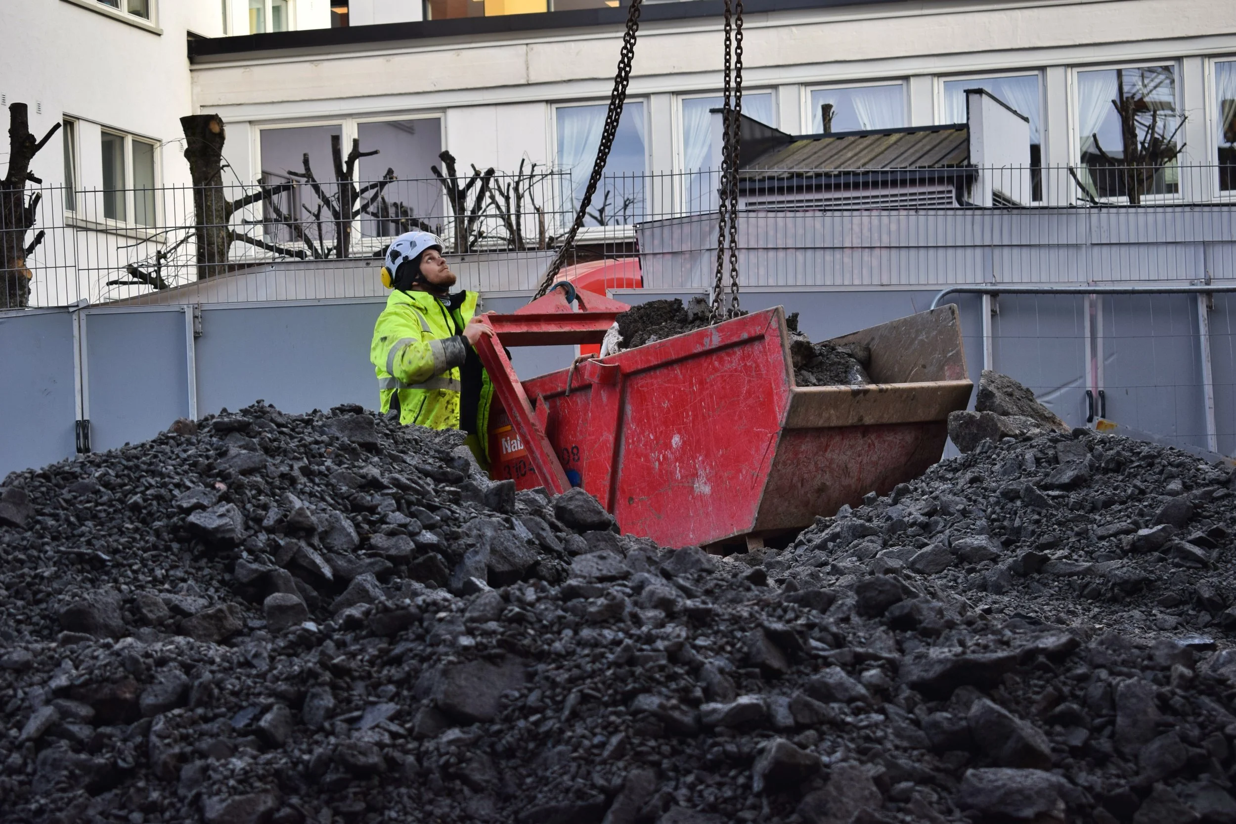 En bygningsarbeider i en gul vest og hjelm står ved en gravemaskin som bumper opp grus eller jord på en byggeplass med hus i bakgrunnen.