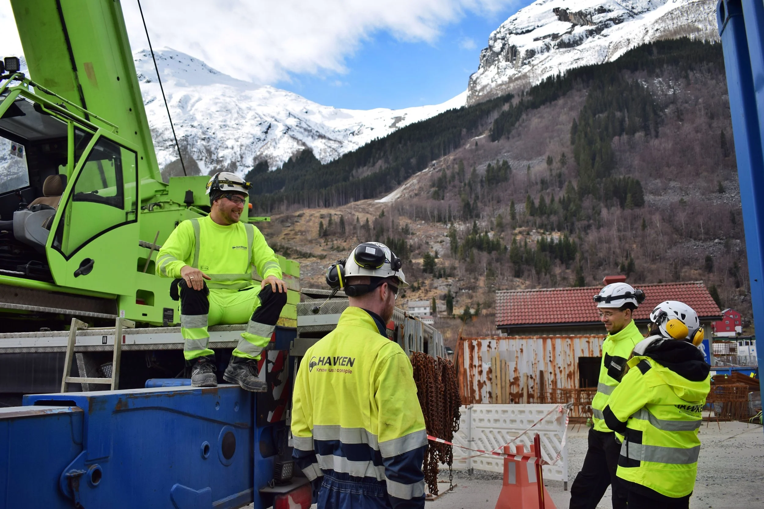 Grupp av bygningsarbeidere i høyt synkende, gule arbeidsklær med hjelm, smilende og snakkende, rundt en stor lastebil med kran, i et område med fjell og fjellhytter i bakgrunnen.
