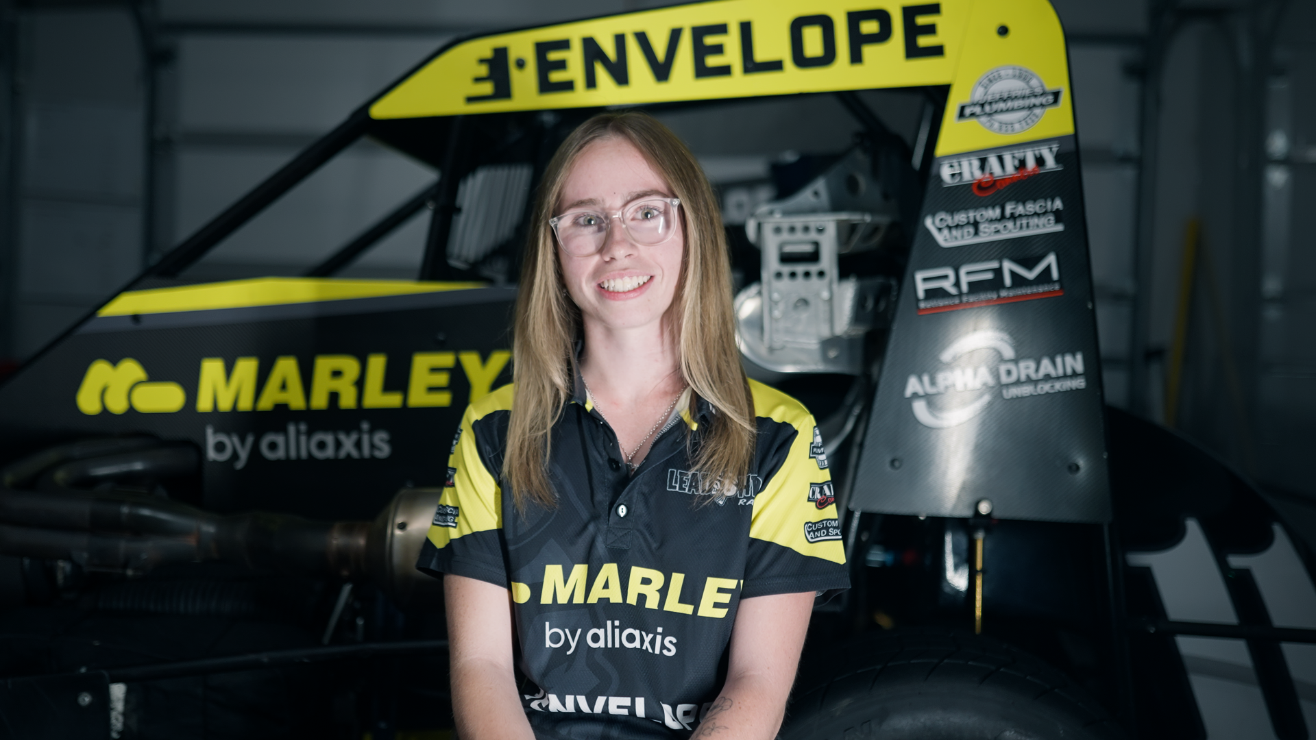 A young woman with glasses and long blonde hair smiling, wearing a yellow and black racing team shirt, sitting in front of a race car in a garage expressing her love for the sport