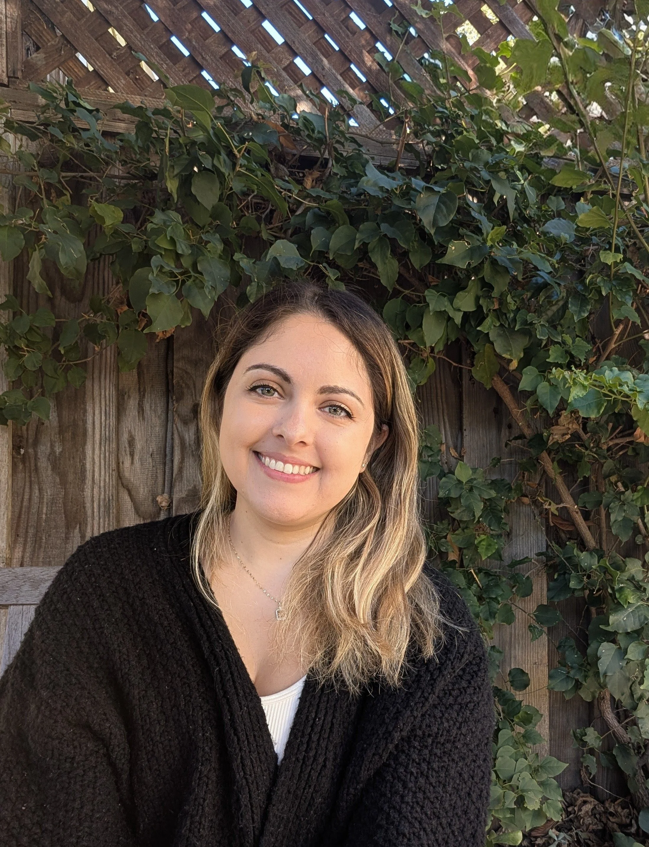 A young woman with shoulder-length wavy blonde hair, wearing a black sweater and a white top, smiling while sitting outdoors in front of a wooden fence covered with green ivy leaves.