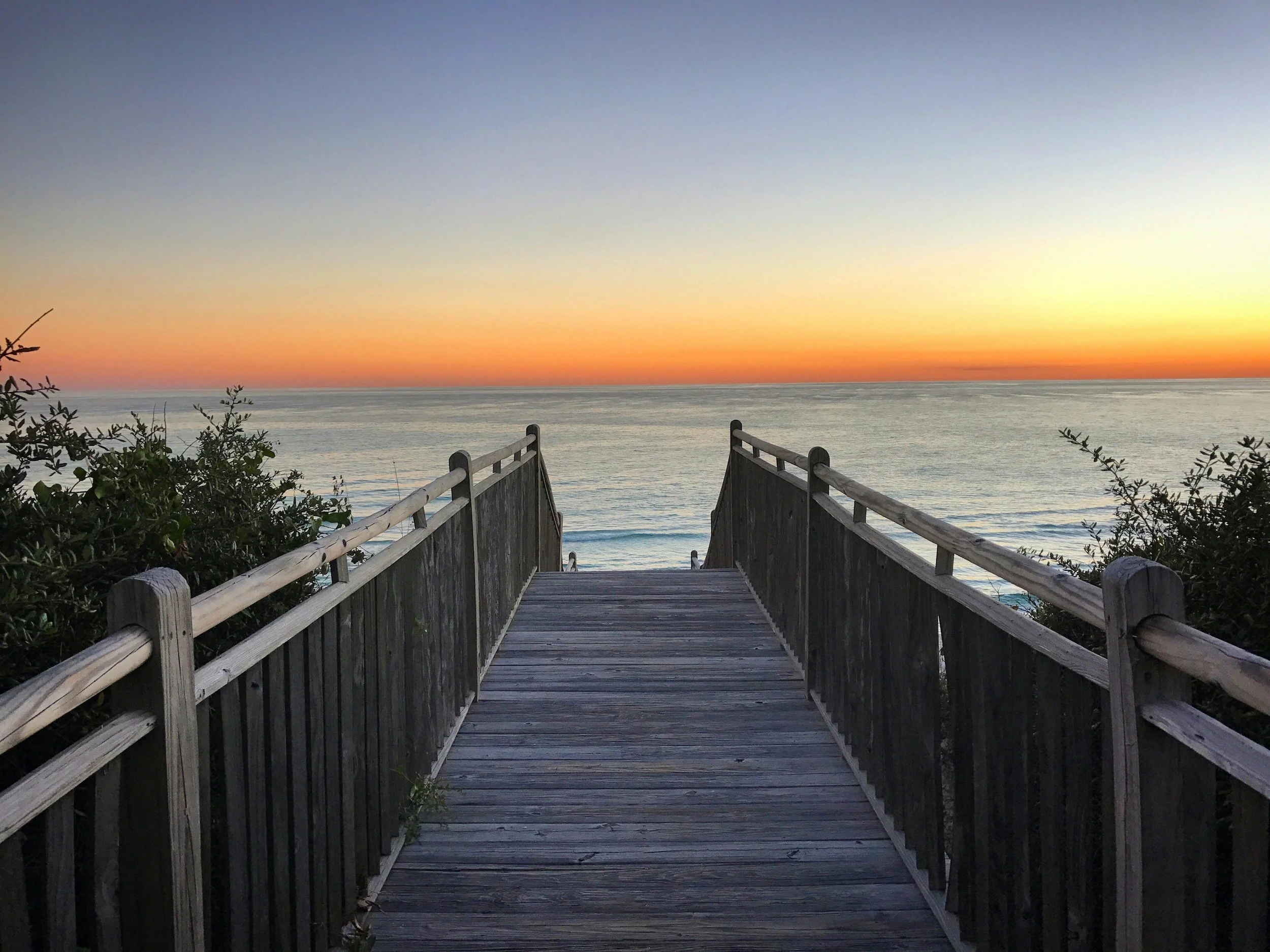A wooden walkway with railings leading to the beach, overlooking an ocean at sunset with a colorful sky.