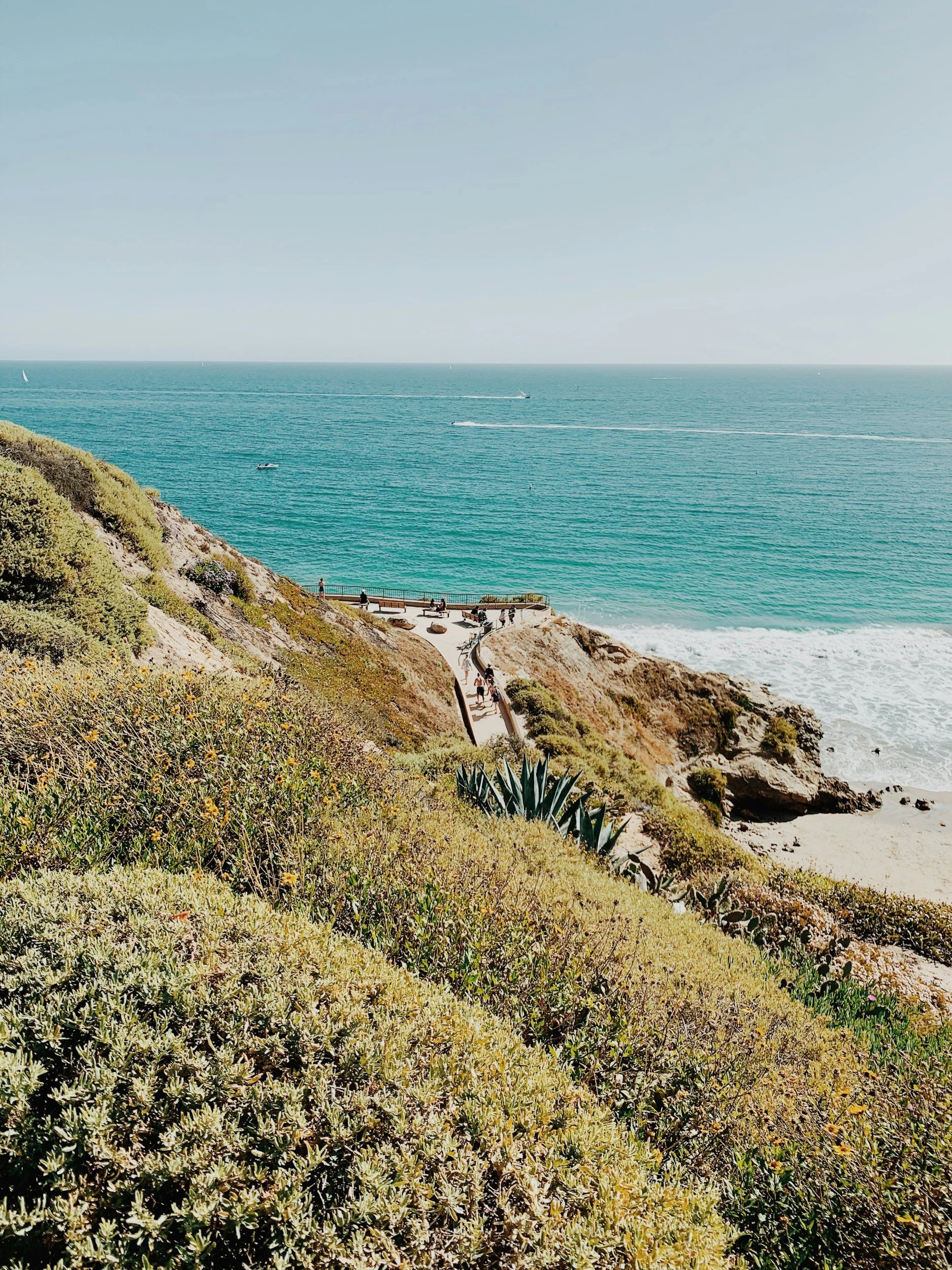 A scenic view of a coastal area with a sandy beach, clear blue ocean, and a paved walkway on a hillside surrounded by green and yellowish bushes, with people walking along the path.
