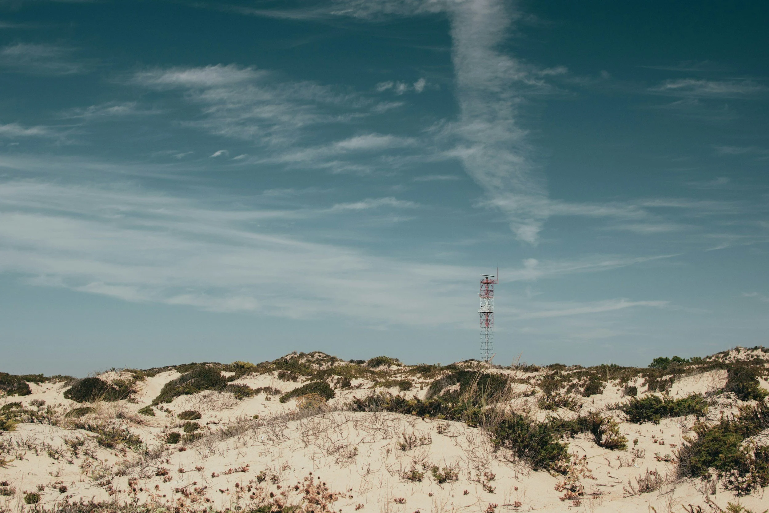Sand dunes with low green shrubs and a red and white communication tower in the distance under a blue sky with wispy clouds.