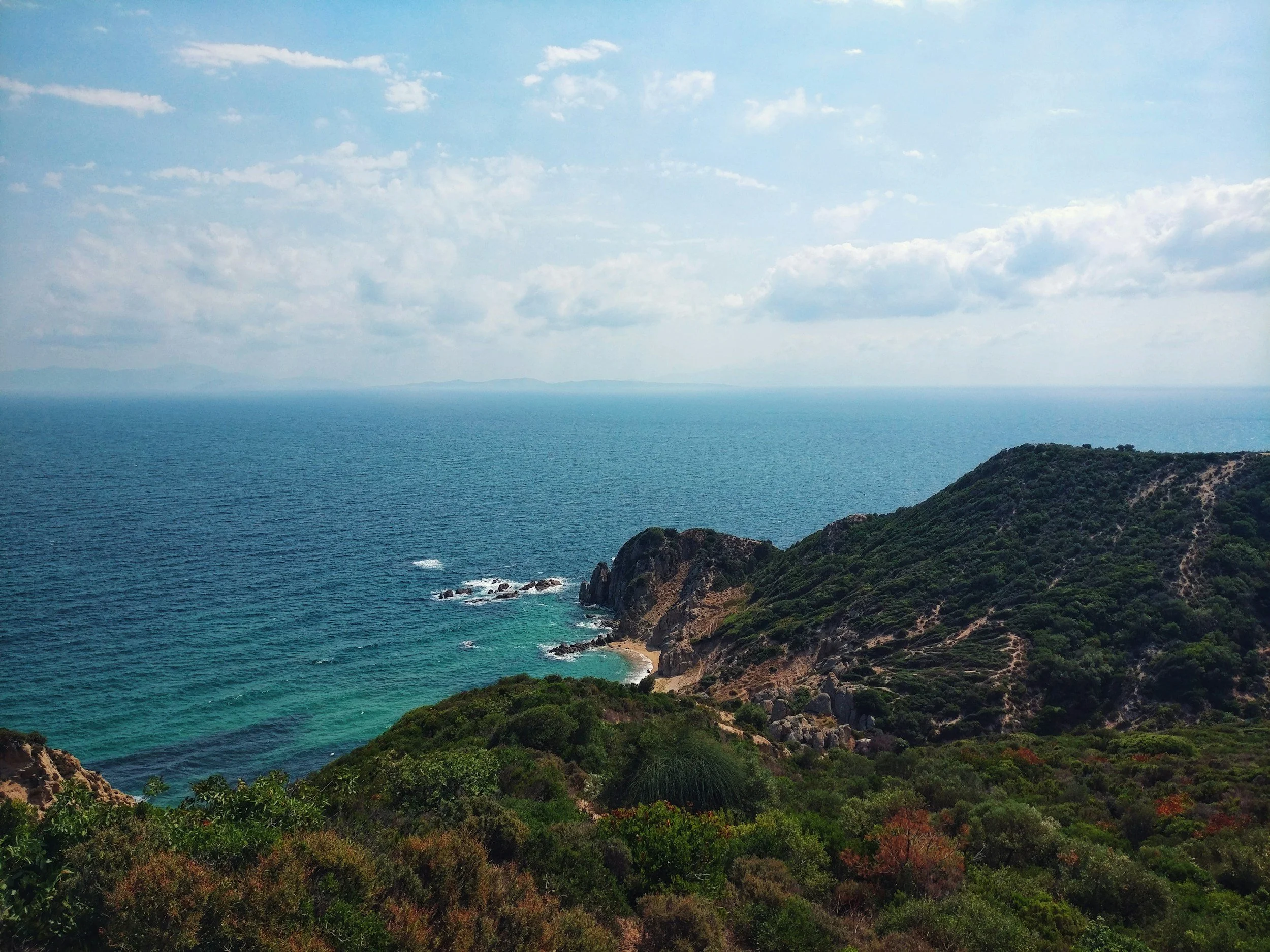 Coastal landscape with ocean, rocky cliffs, and green vegetation under a partly cloudy sky.