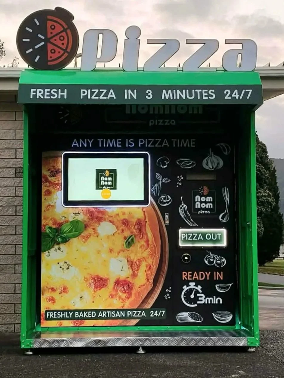 Green pizza vending machine advertising fresh artisan pizza ready in 3 minutes, with a display reading 'PIZZA OUT' and illustrations of pizza ingredients.
