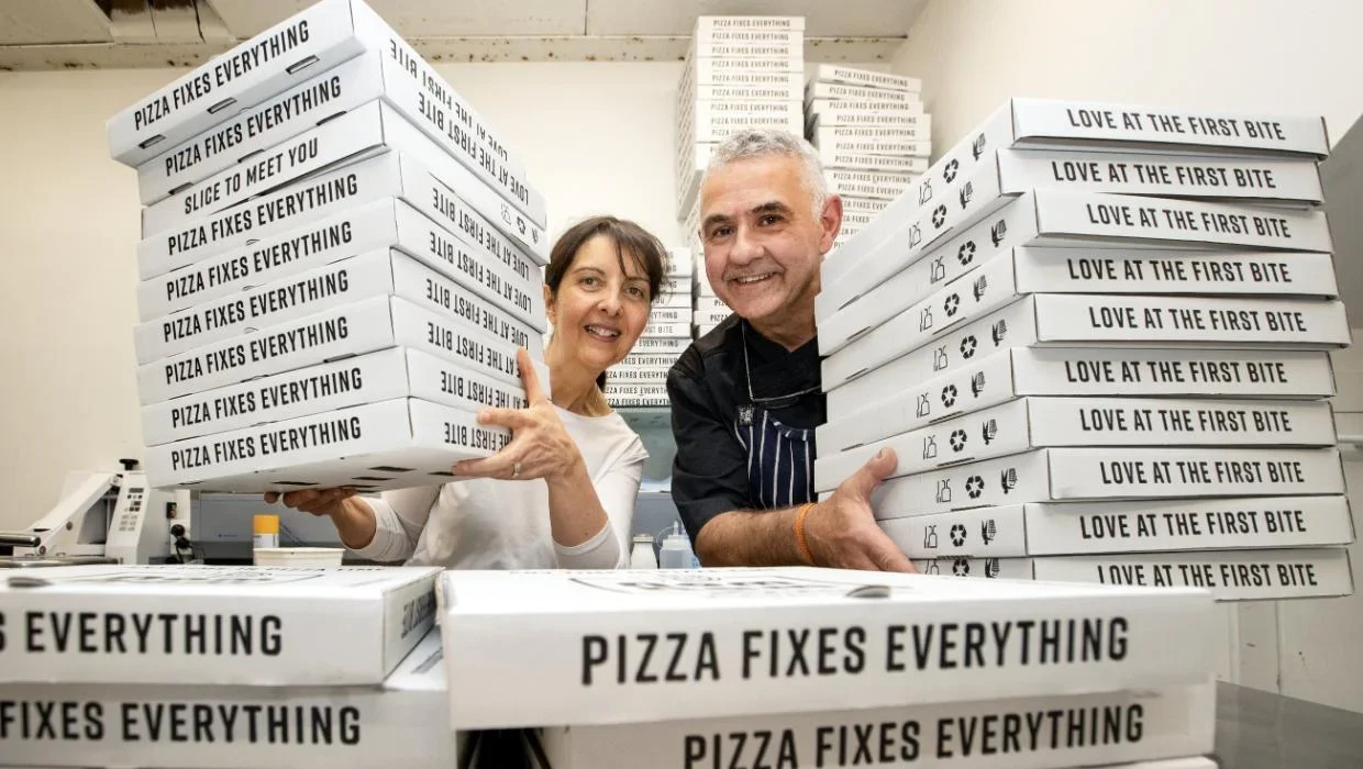 Two people, a woman and a man, stand behind a large pile of pizza boxes in a kitchen or pizzeria. The boxes are labeled with slogans: 'Pizza Fixes Everything,' 'Love at the First Bite,' and 'Slice to Meet You.' The woman is holding several boxes, and both are smiling at the camera.