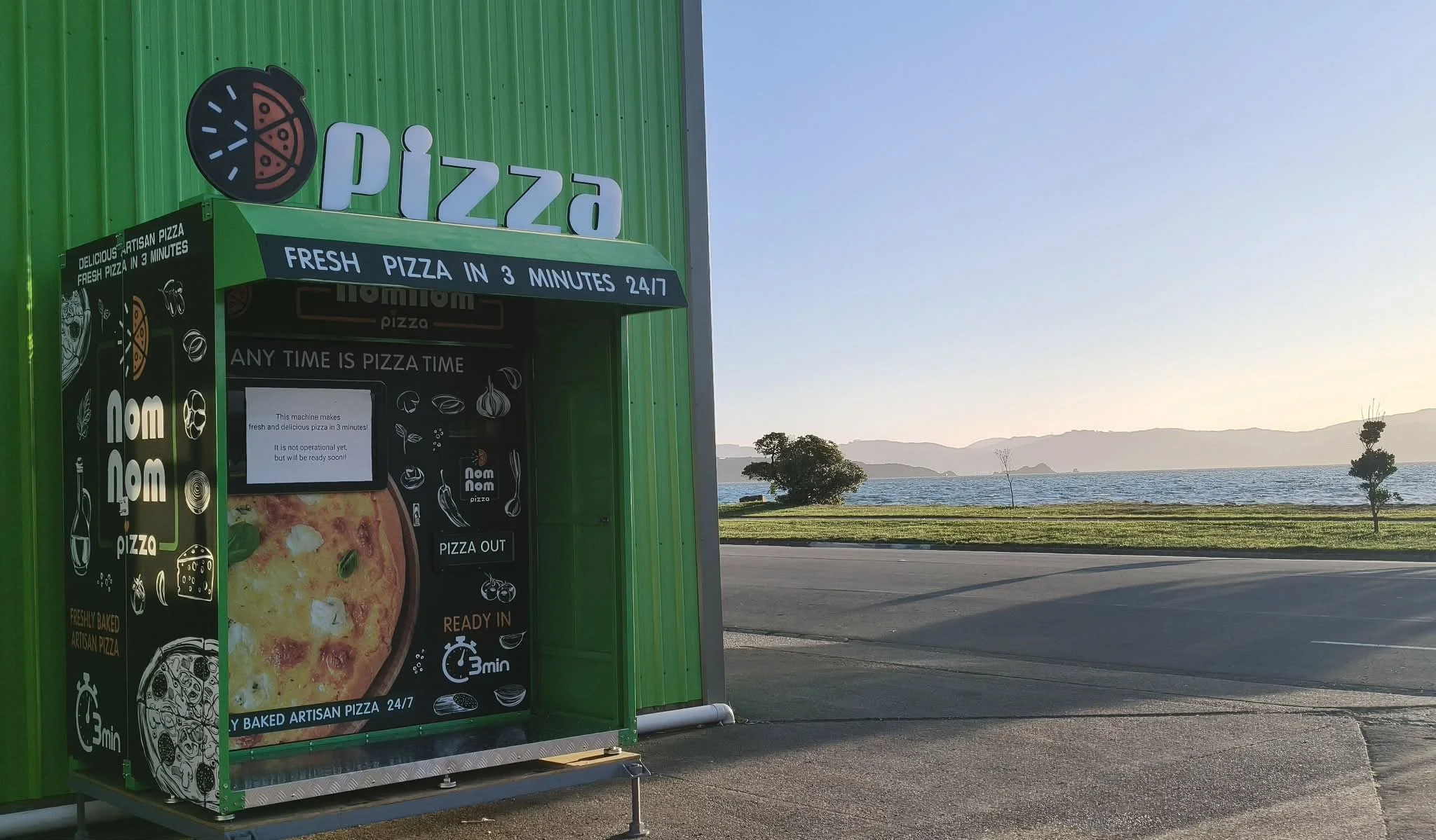 Green pizza vending machine beside a road with a view of a body of water and hills in the background during sunset.