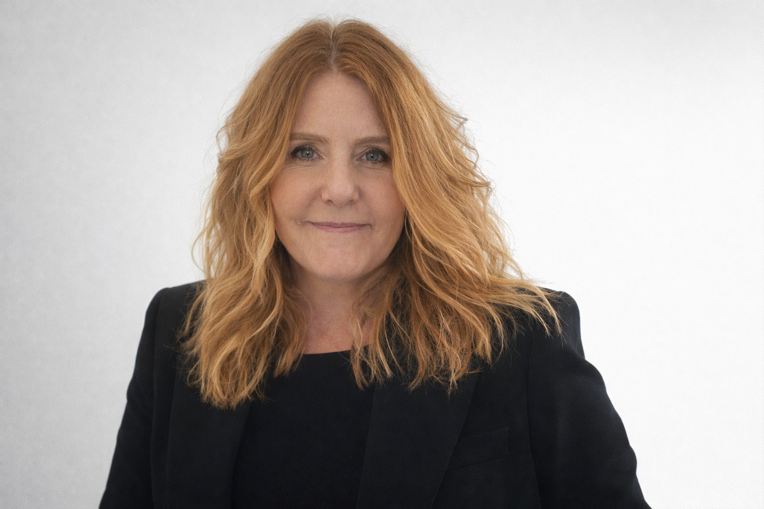 A woman with wavy red hair wearing a black blazer and top, smiling slightly against a plain light background.