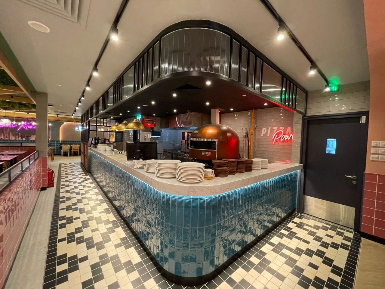 Pizza restaurant counter with pizza oven, stacks of plates, and a neon sign that reads 'Pizza Pur' inside a modern restaurant with checkered tile floors.