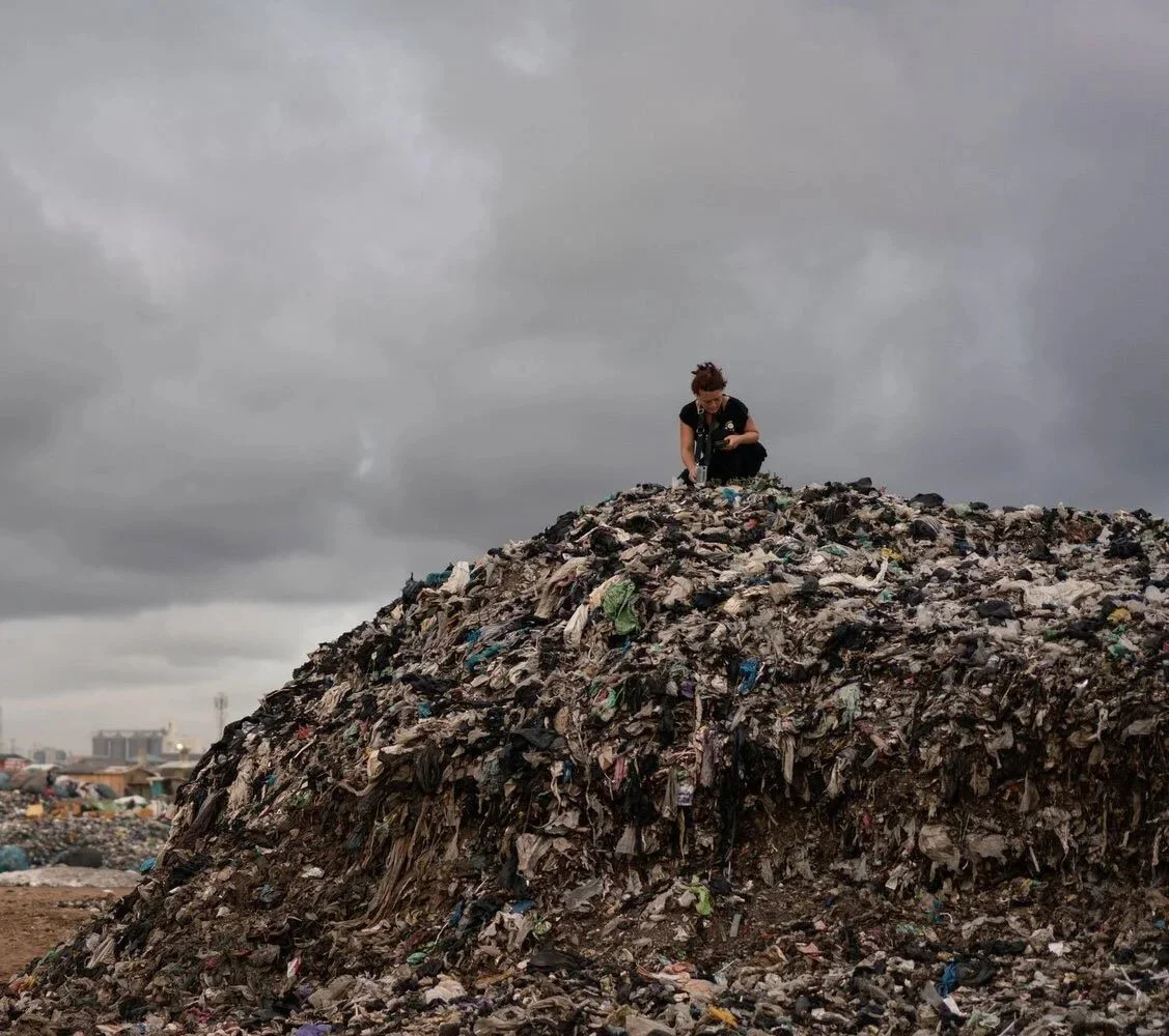 Eine Person sitzt auf einem großen Müllhaufen aus Plastik und Abfällen unter einem bewölkten Himmel.