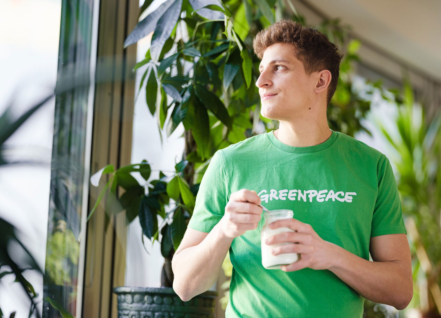 Junge Person in grüner Greenpeace-T-Shirt, die in einer grünen Pflanzenumgebung eine Milch im Glas hält und mit einem Löffel isst, lächelnd und nachdenklich schaubar.