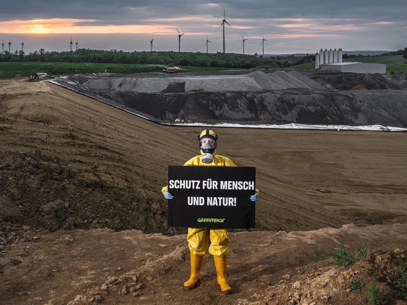 Eine Person in Schutzkleidung trägt ein Schild mit der Aufschrift 'Schutz für Mensch und Natur!' vor einer industriellen Abbaumine mit umfangreichen Anbauanlagen. Im Hintergrund sind Windräder auf einem Hügel zu sehen.