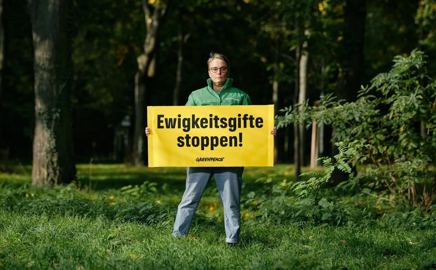 Eine Frau steht im Grünen, hält ein gelbes Plakat mit der Aufschrift "Ewigkeitgifte stoppen!" und trägt eine grüne Jacke, vor einem Wald.
