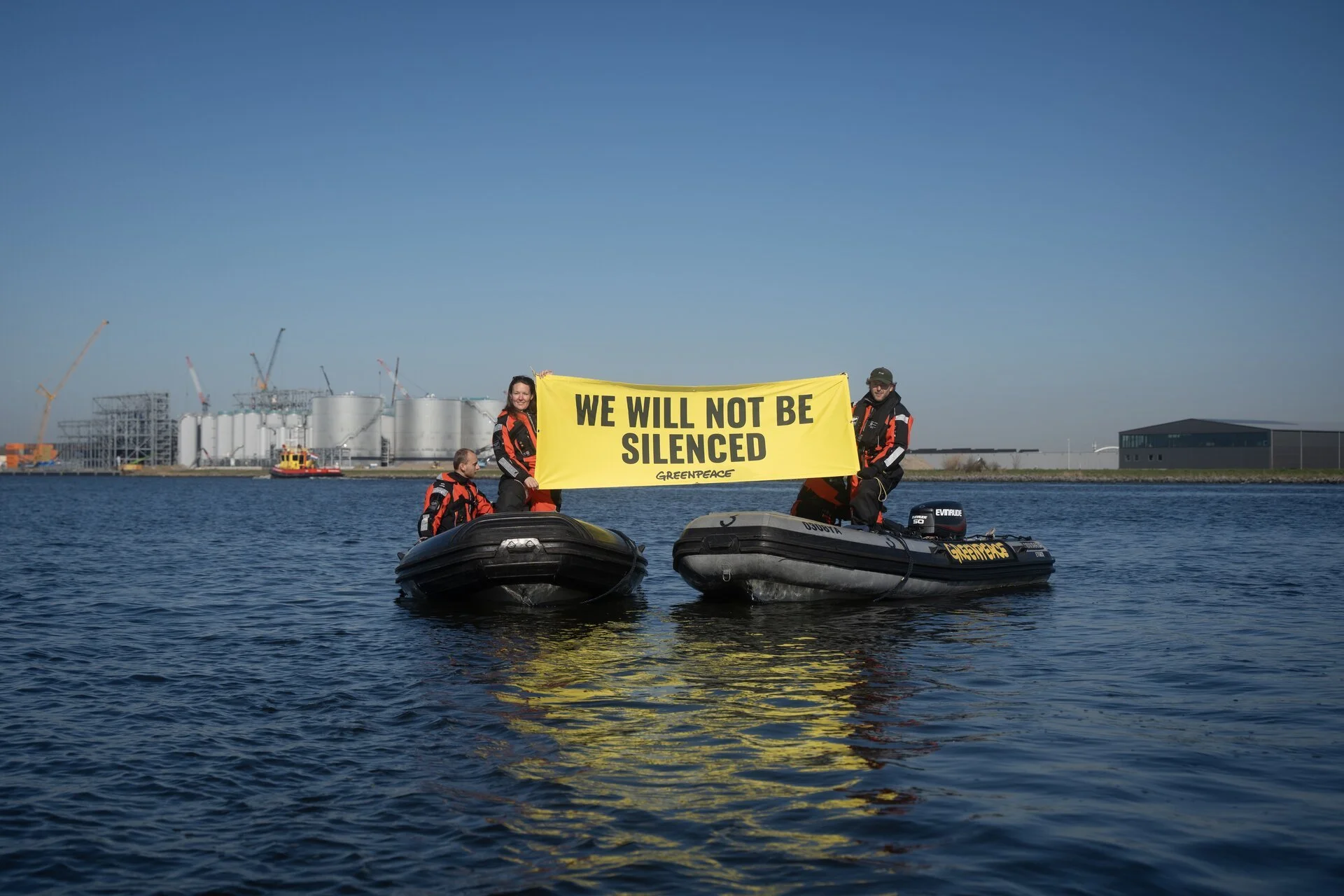 Zwei Menschen auf einem Boot auf dem Wasser halten ein gelbes Banner mit der Aufschrift ‚We will not be silenced‘. Im Hintergrund sind Fabrikanlagen und Baukräne sichtbar.