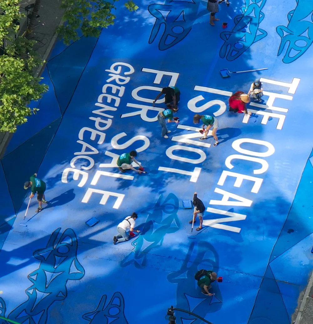 Mehrere Personen beim Malen eines großen blauen Wandbildes mit weißen und schwarzen Schriftzügen, das den Titel 'THE OCEAN IS NOT GREED, PEACE' trägt, während sie verschiedene Abschnitte der Wand mit Pinseln und anderen Werkzeugen gestalten.
