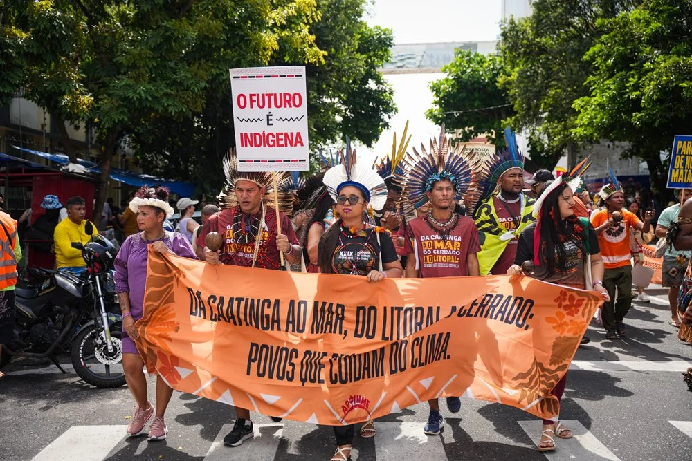 Menschen eines indigenen Stammes protestieren auf einer Straße, halten ein Banner mit portugiesischem Text und tragen traditionelle Kleidung sowie Kopfschmuck aus Federn.