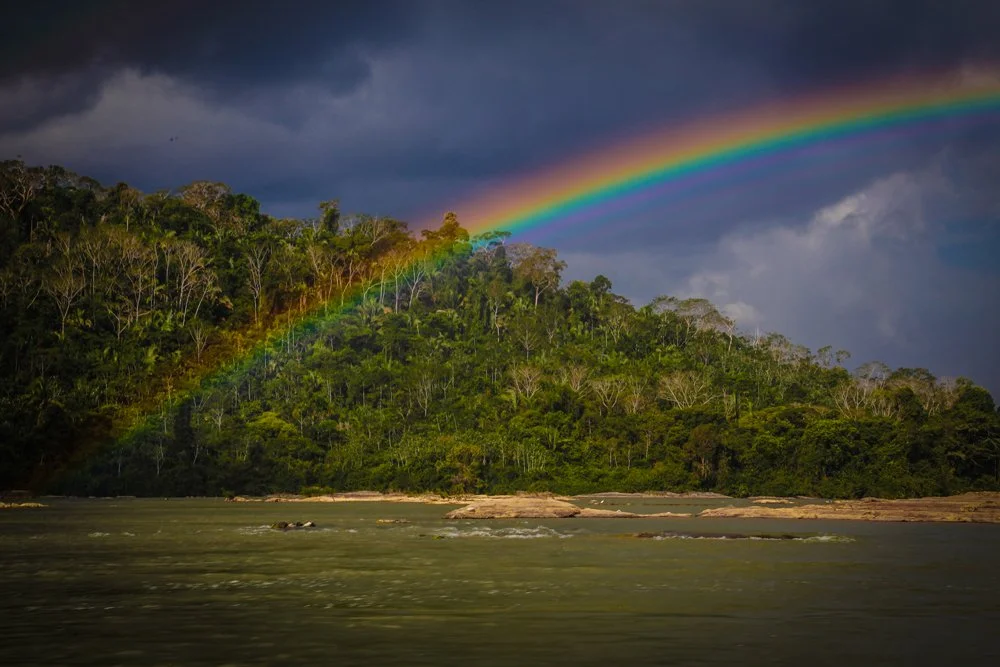 Regenbogen über einem bewaldeten Hügel am Fluss unter dunklen Wolken.