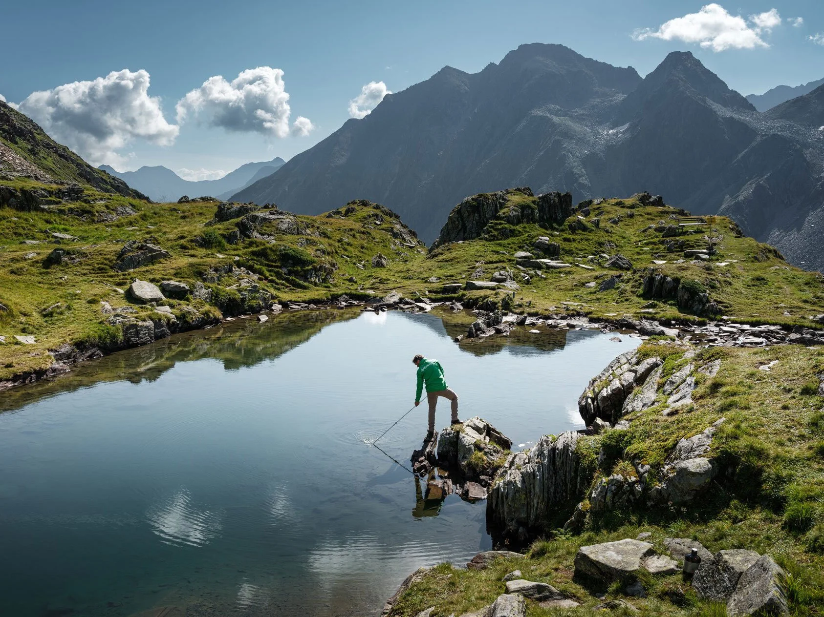 Ein Mensch steht in einem kleinen See in den Bergen und hält ein Angelrute, umgeben von grünen Hügeln und felsigen Landschaften, mit hohen Bergen im Hintergrund und einem blauen Himmel mit wenigen Wolken.