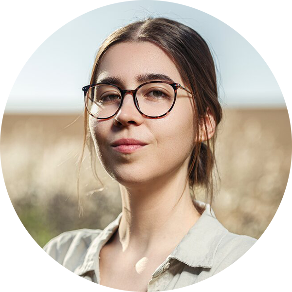 Junge Frau mit Brille und beige Hemd steht im Freien, Himmel im Hintergrund.