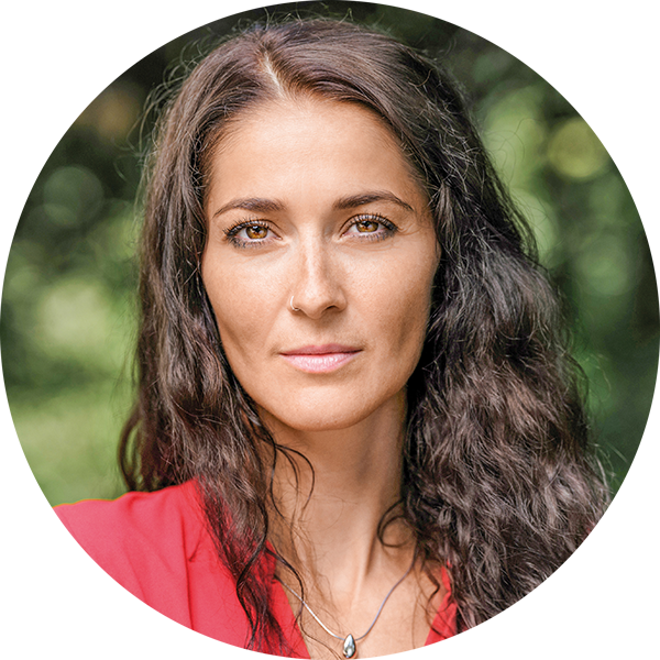 Nahaufnahme einer jungen Frau mit braunen, lockigen Haaren, die ein rotes Top trägt, vor grünem, unscharfem Hintergrund.