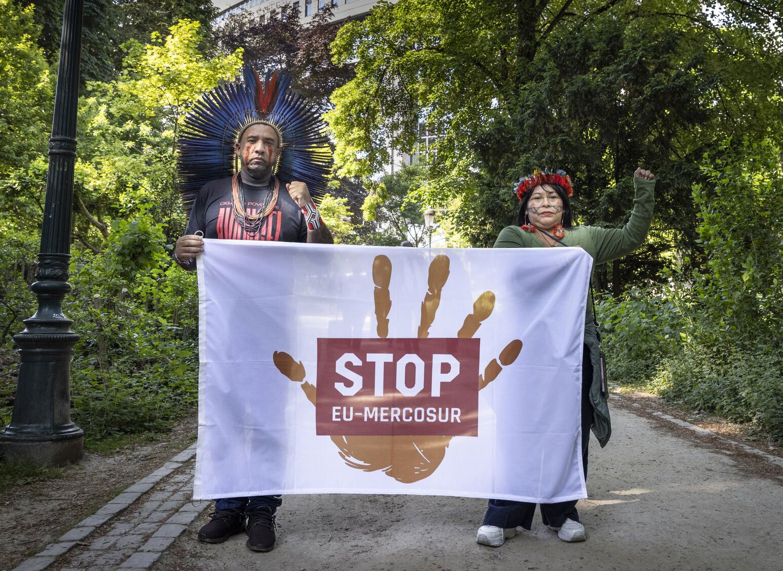 Zwei Demonstranten halten ein Banner mit der Aufschrift 'STOP EU-MERCOSUR'. Der Mann auf der linken Seite trägt ein Fell-Kopfschmuck und ein T-Shirt mit roten und schwarzen Mustern. Die Frau auf der rechten Seite trägt eine Blumenkrone und Brillen. Sie stehen in einem bewaldeten Park, umgeben von grünen Bäumen.
