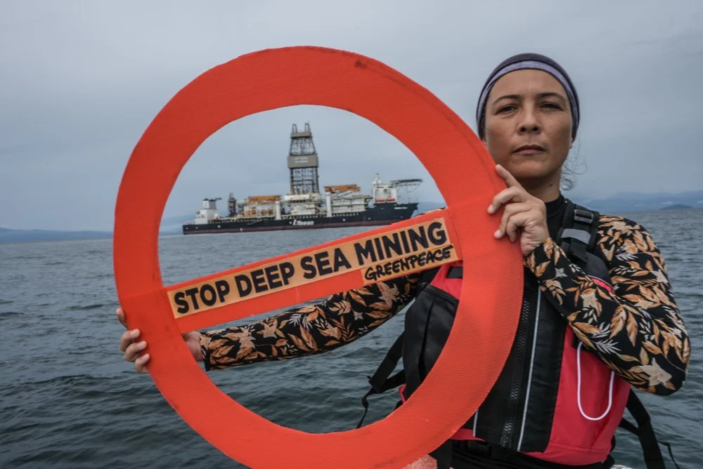 Frau hält ein rotes Symbol mit der Aufschrift 'Stop Deep Sea Mining Greenpeace' vor dem Meer, im Hintergrund ein Schiff, Greenpeace-Aktion gegen Tiefseebergbau.