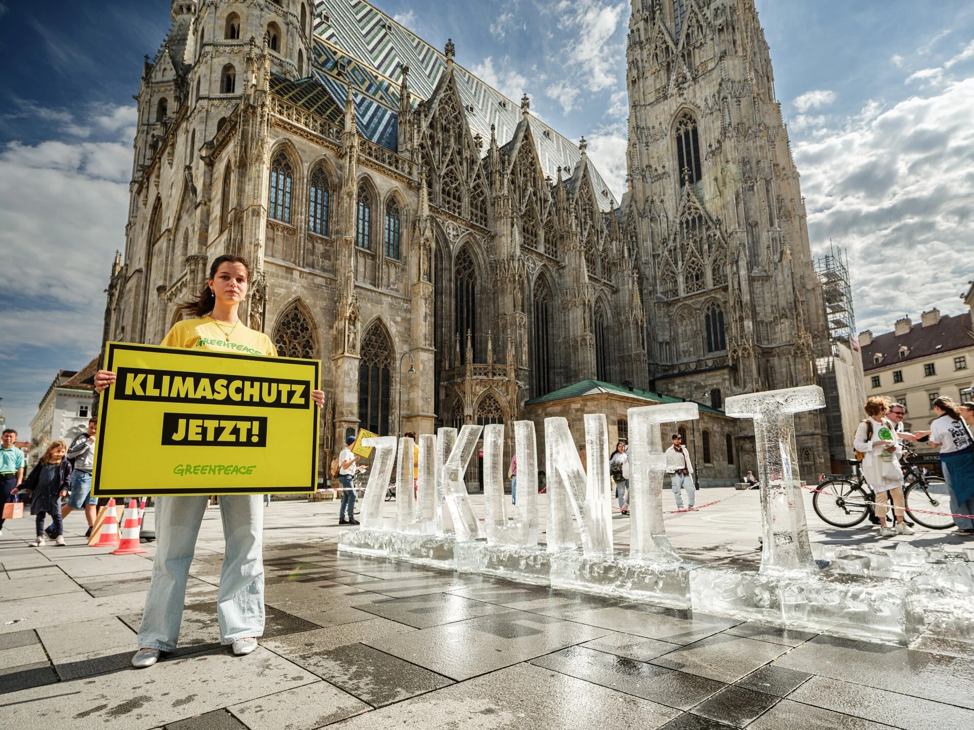 Eine Frau hält ein Schild mit der Aufschrift ‚Klimaschutz jetzt!‘ vor der Kathedrale in einer belebten Stadt, umgeben von anderen Menschen und Eisskulpturen im öffentlichen Raum.