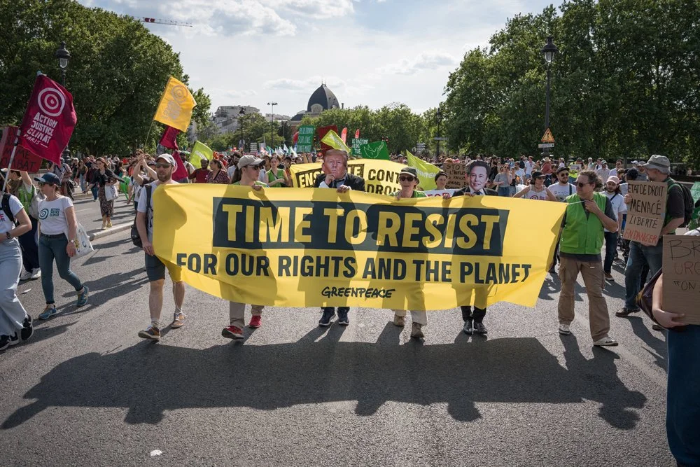 Protestmarsch mit Menschen, die Schilder und Banner tragen, darunter ein großes gelbes Banner mit der Aufschrift "TIME TO RESIST FOR OUR RIGHTS AND THE PLANET"