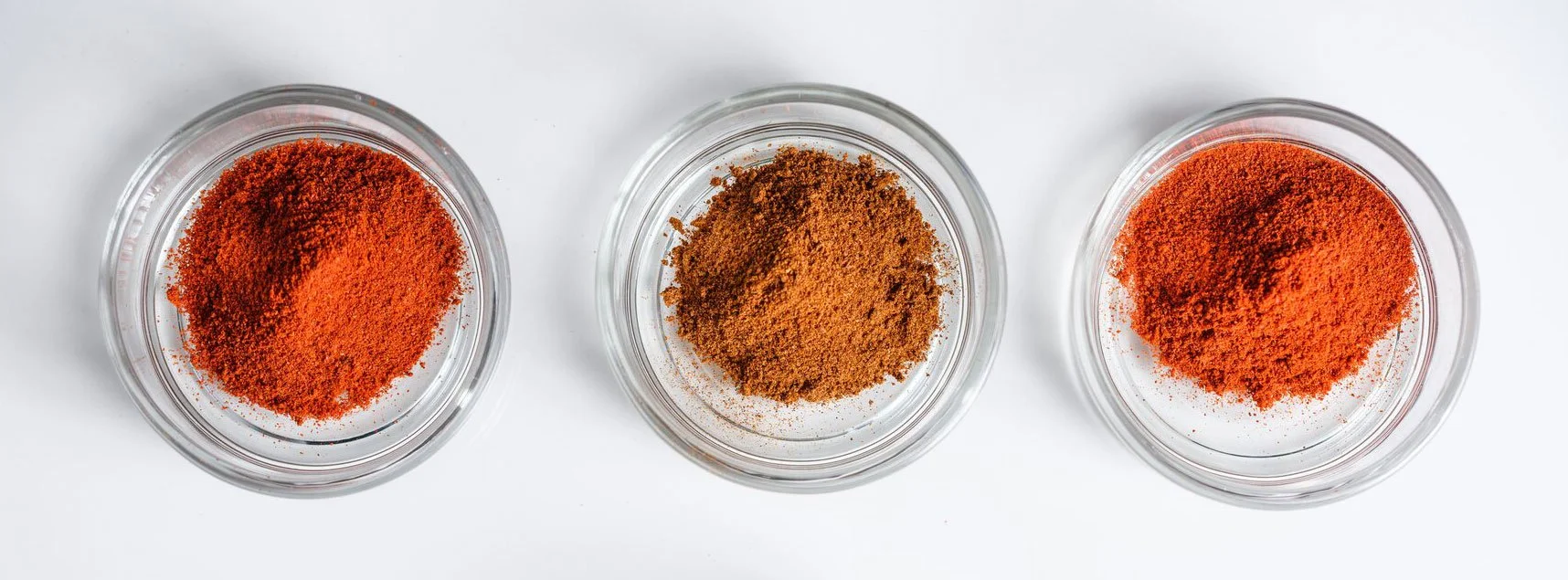 Drei kleine Glas-Schüsseln auf weißem Hintergrund, jeweils gefüllt mit rotem, braunem und orangem Gewürzpulver.