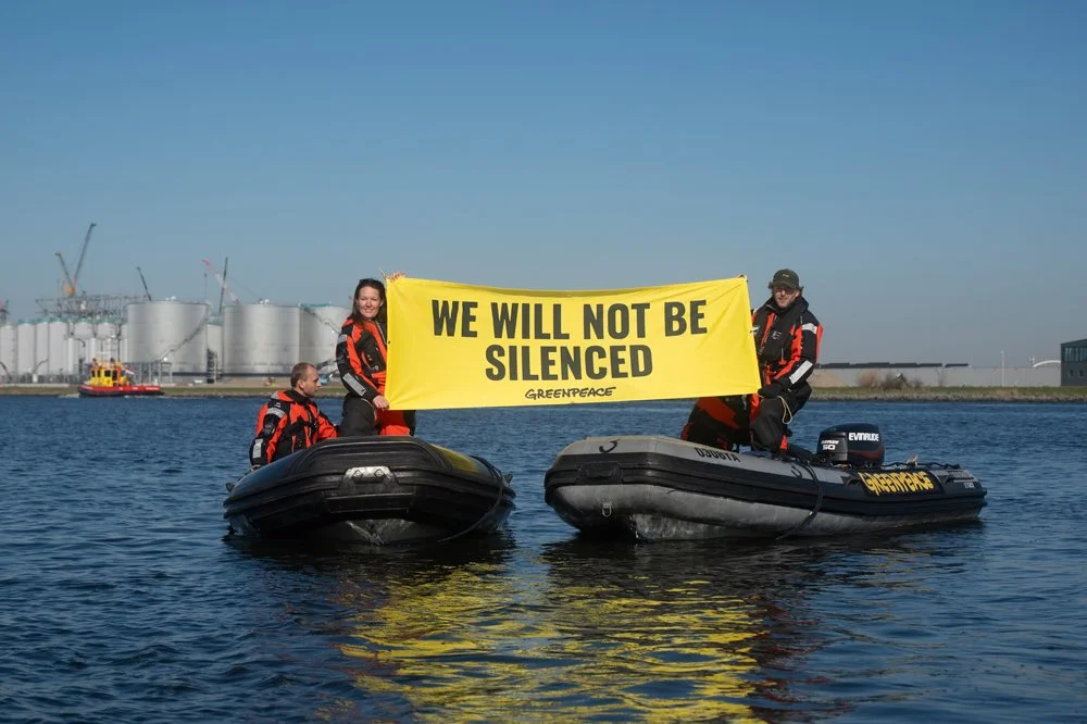 Menschen auf zwei Booten halten ein gelbes Banner mit der Aufschrift "We will not be silenced" bei Greenpeace, im Hintergrund ein Industriegebiet mit Tanks und Kränen.
