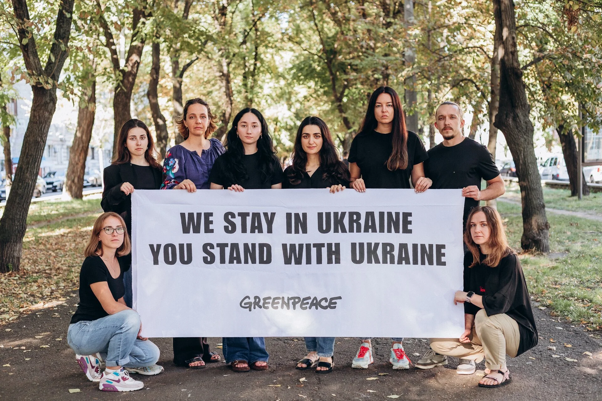 Gruppe von Menschen, die in einem Park stehen und eine Greenpeace-Fahne halten, mit einem Baumhintergrund, während sie für die Unterstützung der Ukraine demonstrieren.