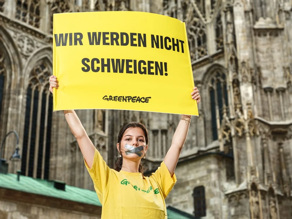 Junge Frau protestiert mit einem gelben Schild, das 'Wir werden nicht schweigen!' und Greenpeace geschrieben steht, und trägt Tuch vor dem Mund, vor einer alten Kathedrale.