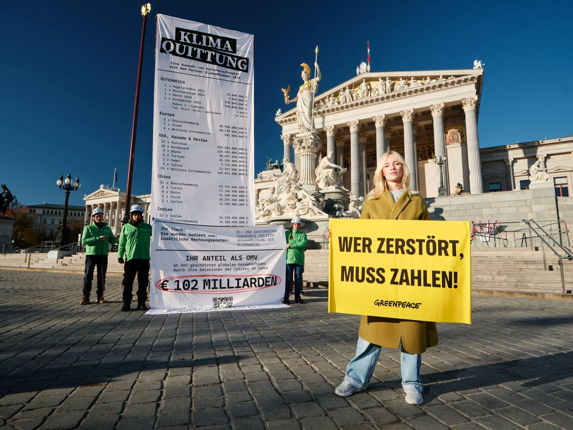 Junge Frau hält ein gelbes Schild mit der Aufschrift 'WER ZERSTÖRT, MUSS ZAHLEN!'. Im Hintergrund stehen weitere Demonstranten und ein großes Banner, das Klimawandel und Umweltkosten thematisiert, vor dem Parlament in Wien.
