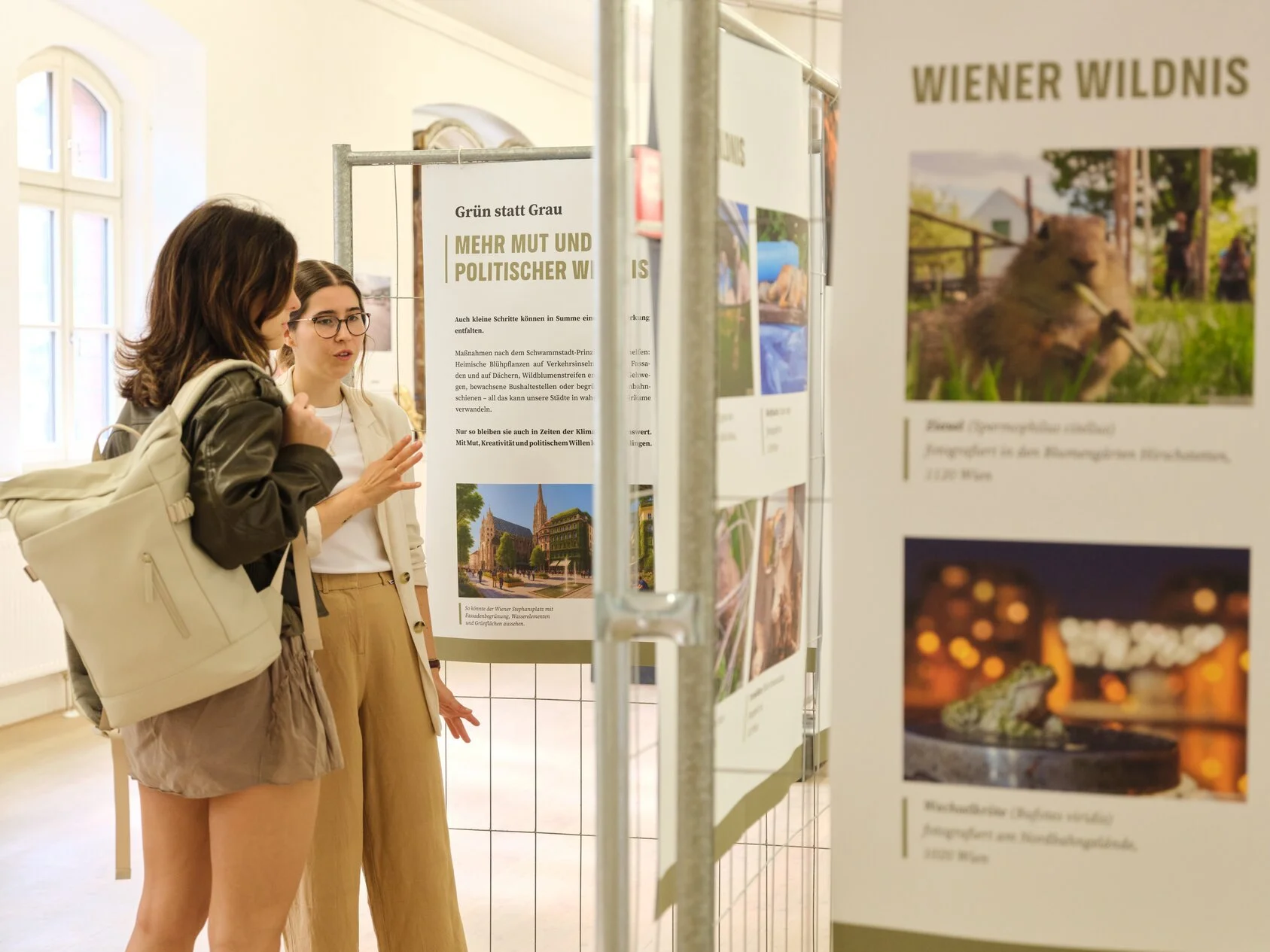 Zwei junge Frauen diskutieren vor einem Ausstellungsdisplay mit Fotos und Text im Innenraum eines Museums oder einer Galerie.