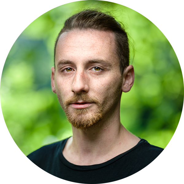 Porträt eines jungen Mannes mit hellbraunem Haar und Bart vor einem grünen, bewachsenen Hintergrund.