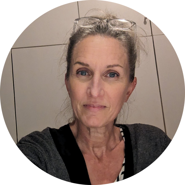 Ein Porträt einer Frau mit blauen Augen und grauem Haar, die eine Brille auf dem Kopf trägt, vor einer weißen Wand.