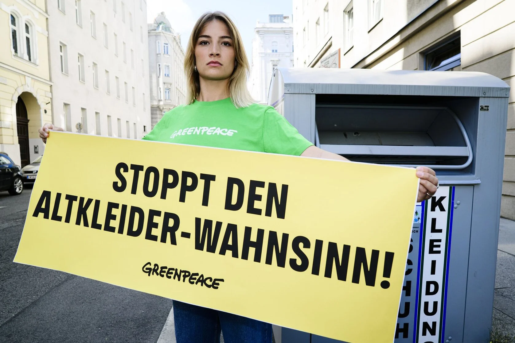 Eine junge Frau trägt ein grünes Greenpeace-T-Shirt und hält ein gelbes Banner mit der Aufschrift "Stoppt den Altkleider-Wahnsinn!" vor einem Postkästchen auf einer Stadtstraße.