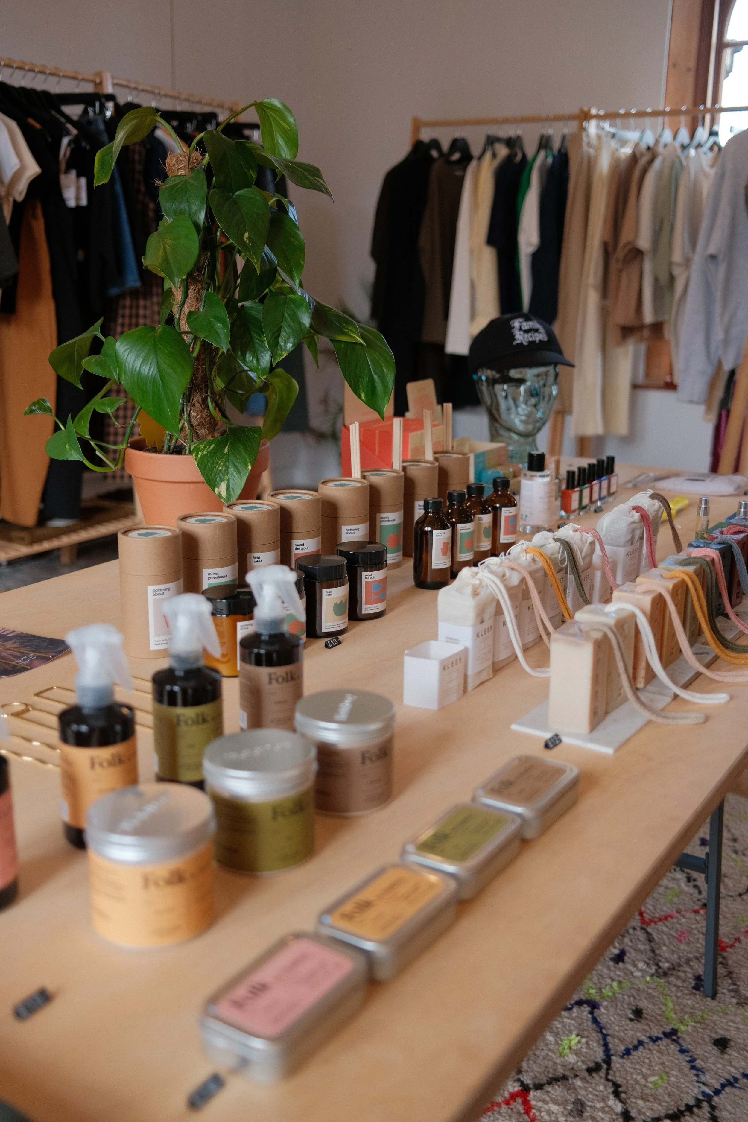 A store display with a potted green plant, various skincare products in jars and bottles, makeup palettes, and small bags on a wooden table. Clothing hangs on racks in the background.