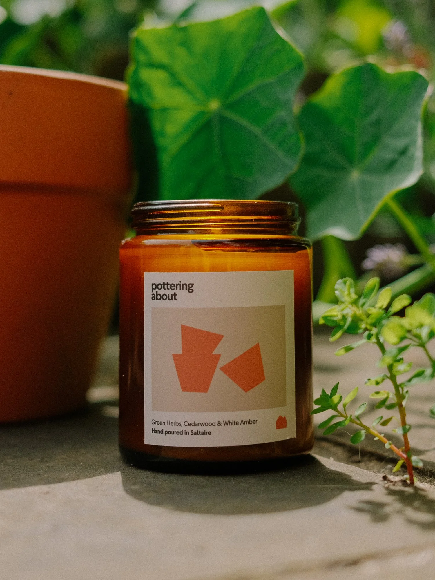 A jar of Pottering About hand-poured candle with a label indicating it contains green herbs, cedarwood, and white amber, placed on a surface with green plants in the background.