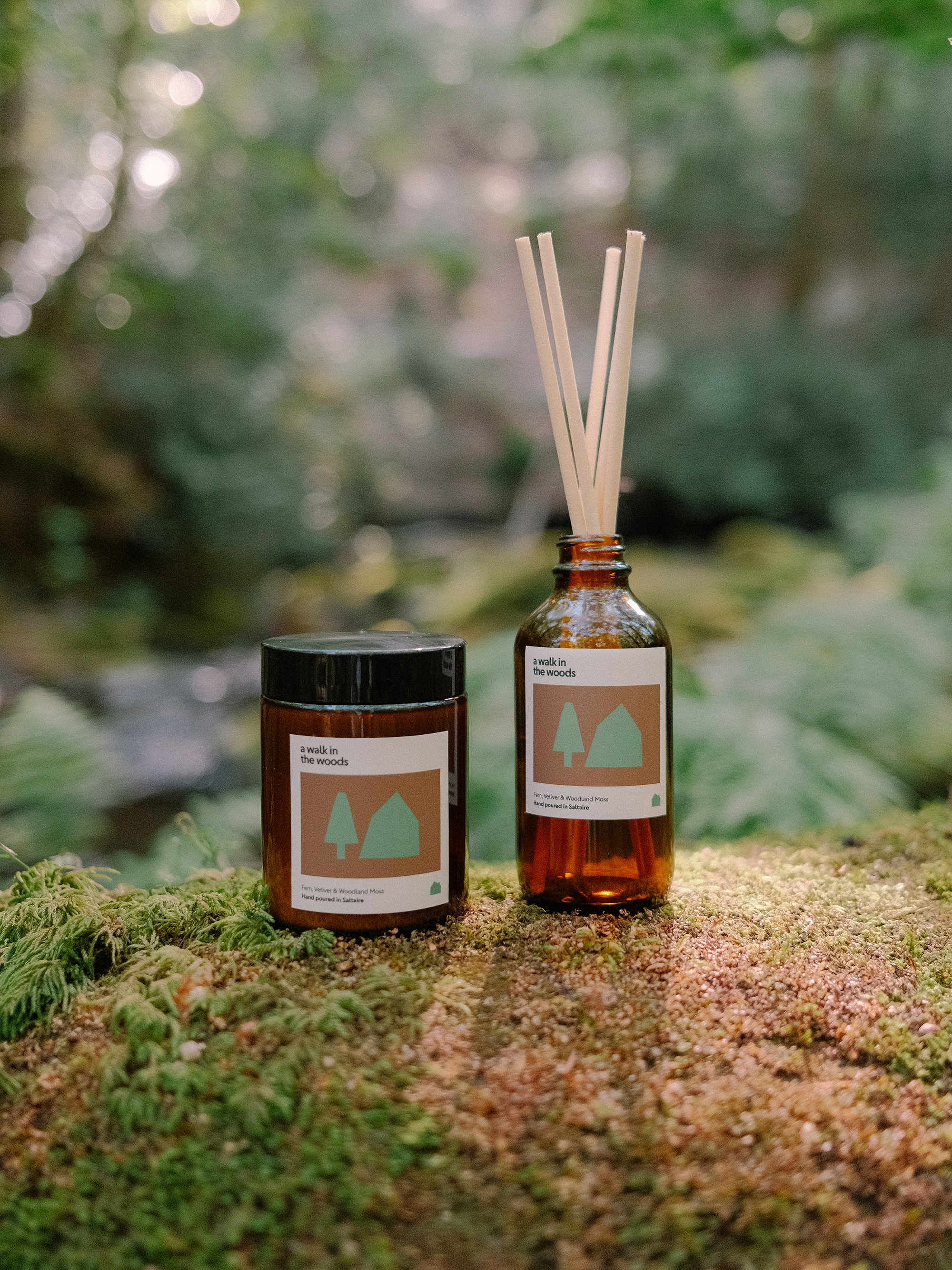 Two amber glass bottles with labels that read "a walk in the woods" and depict stylized trees and mountain shapes. One bottle has a black lid and the other has reed diffuser sticks. The bottles are placed on a mossy surface with a blurred green forest background.
