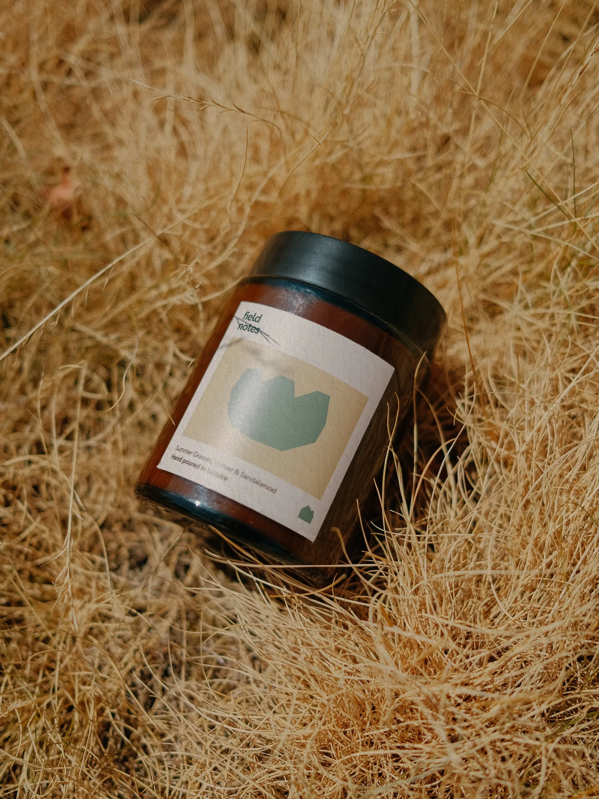 A brown glass jar with a black lid lying on dry, yellowish grass. The jar has a label with the word 'field' and a green mountain-like design.