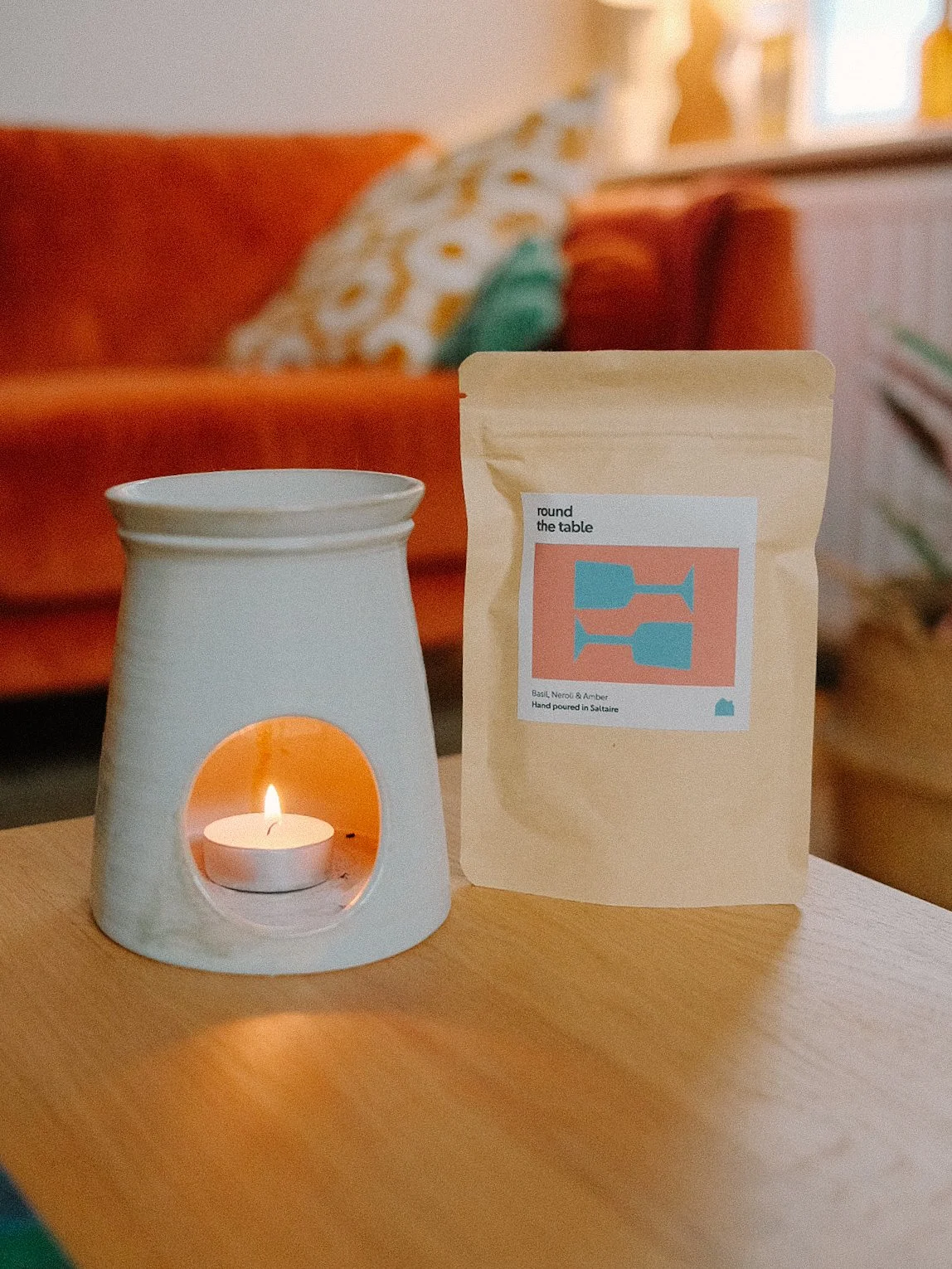 A white candle warmer with a lit tea light candle inside, placed on a wooden surface next to a beige bag of coffee or tea labeled "round the table" with a stylized label design featuring blue and coral shapes, in a cozy room with a blurred red sofa and decorative pillow in the background.