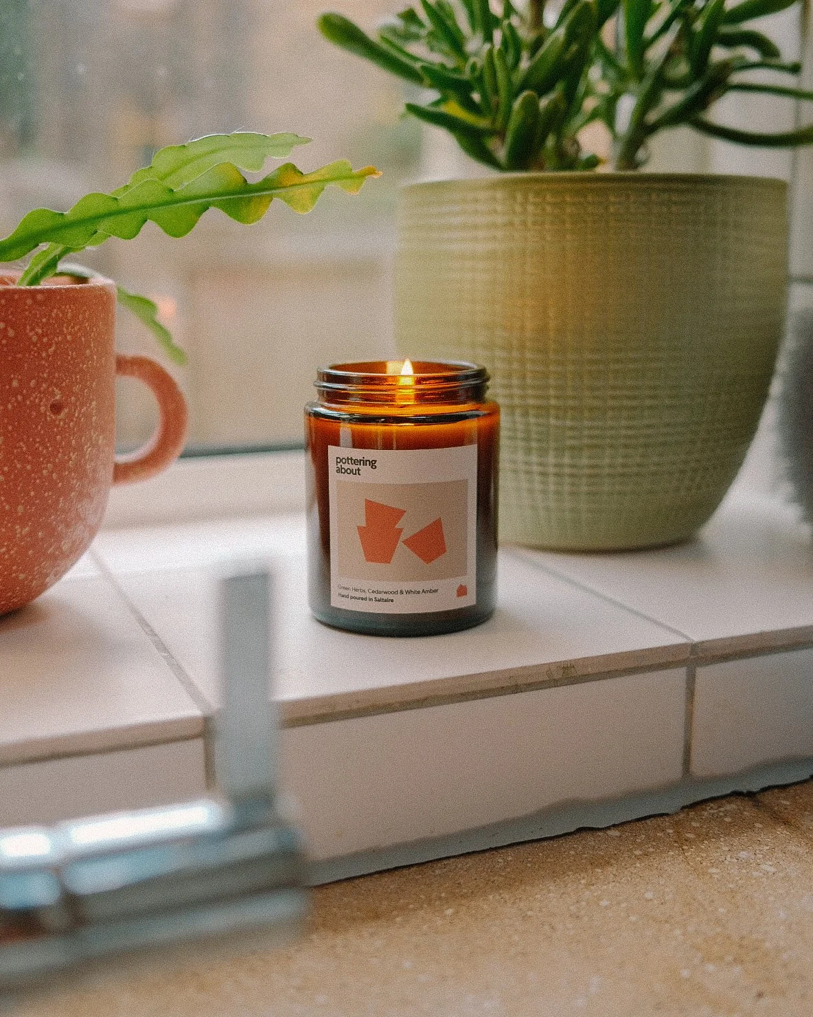 A lit candle in a brown glass jar with a white label, placed on a white tiled surface between two potted plants. One plant has green, wavy leaves in a pink speckled mug, the other has long, thin leaves in a large, textured green pot. The background shows a blurred window.