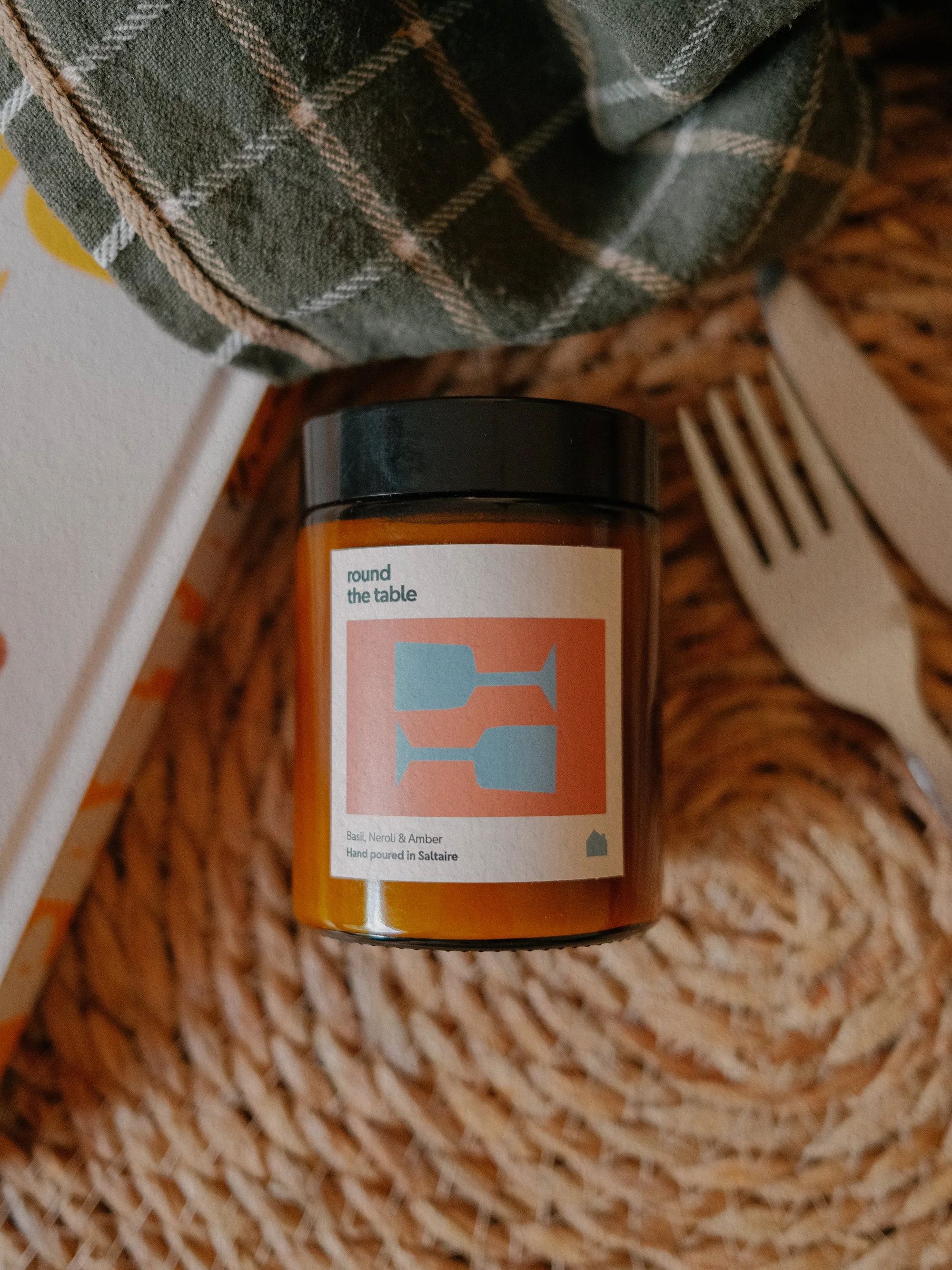A brown glass jar of hand poured candle with a label that reads 'round the table' and contains basil, neroli, and amber scent. The jar is placed on a woven basket surface with a gray plaid cloth and a white fork nearby.