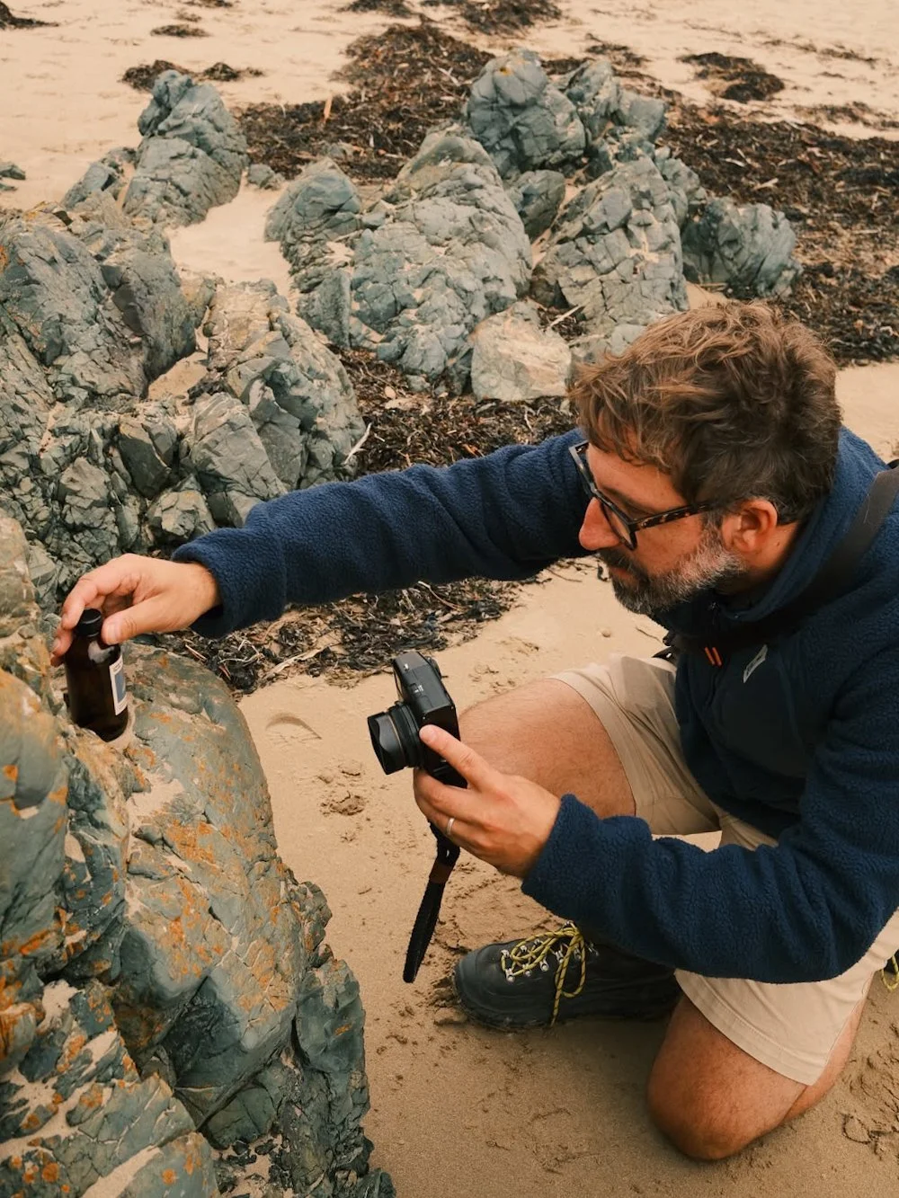 Man kneeling on a sandy beach, photographing rocks with a camera and holding a small bottle, possibly collecting or examining something near the rocks.