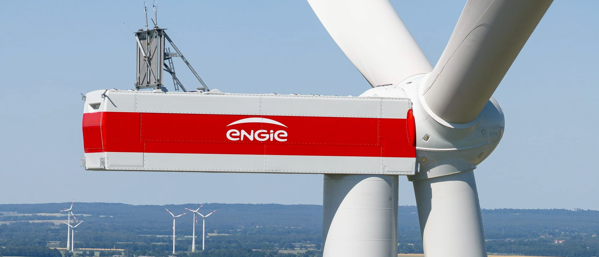 Großer Windkraftgenerator mit einem Rot-weißen Gehäuse und der Aufschrift 'ENGIE' auf einer grünen Wiese, im Hintergrund mehrere kleinere Windräder unter einem blauen Himmel.